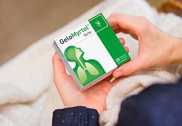 Frau mit Erkältung hält eine Packung GeloMyrtol® forte in der Hand