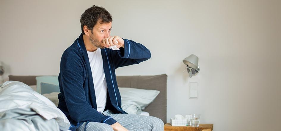 Ein Mann sitzt krank im Bett und hustet: Die Dauer einer Erkältung beträgt in der Regel neun Tage.
